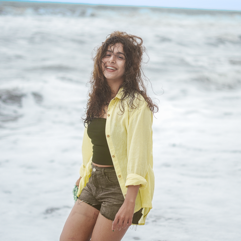 A woman standing in shallow ocean water at the beach, wearing an open light yellow button-up shirt over a dark green top and olive-brown shorts.