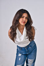 A woman posing in a white sleeveless top and distressed blue wide-leg jeans against a light gray studio background.