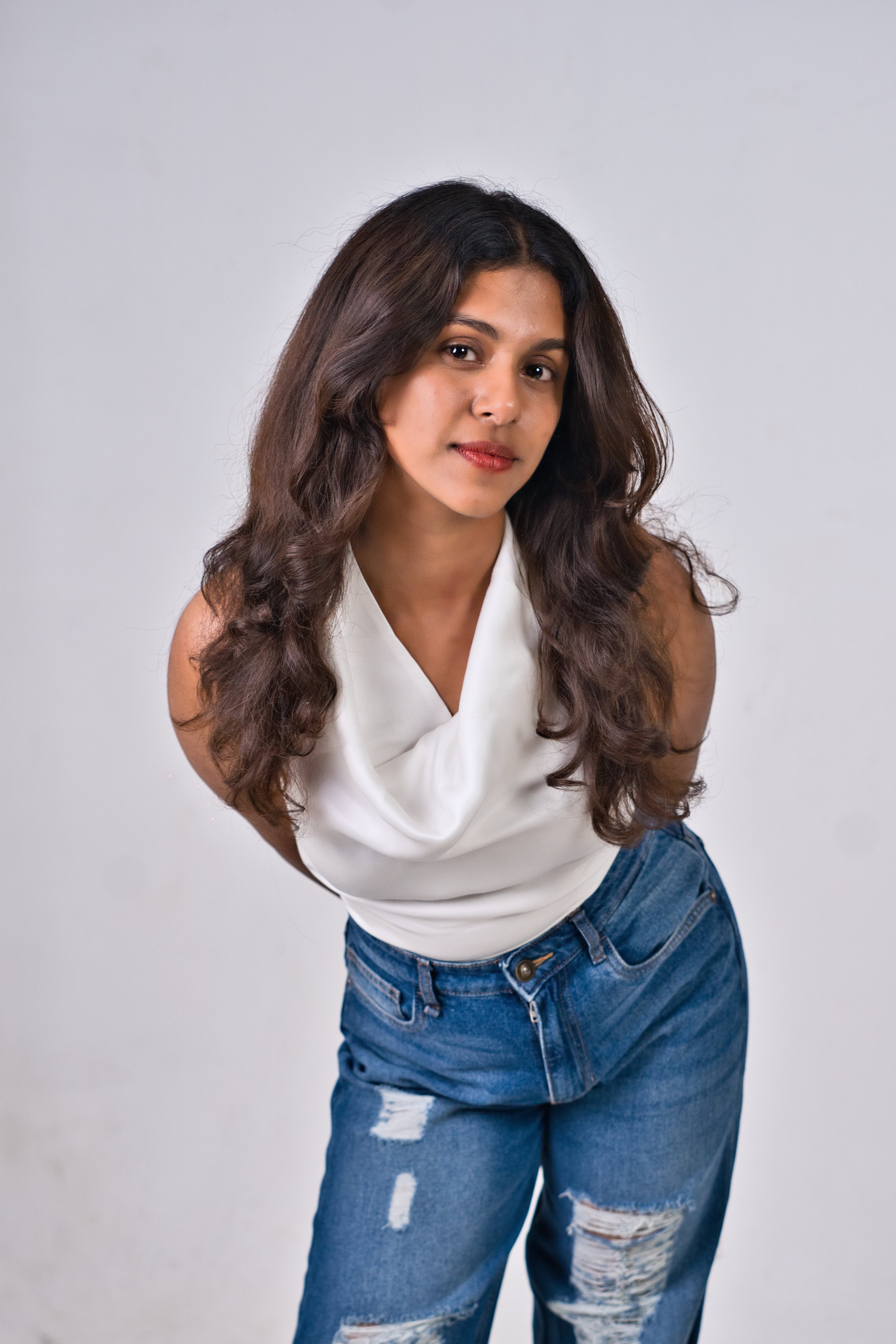 A woman posing in a white sleeveless top and distressed blue wide-leg jeans against a light gray studio background.