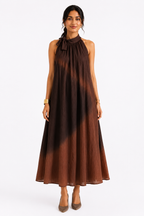 A woman modeling a sleeveless halter-neck maxi dress with a brown ombre tie-dye pattern, standing against a plain white studio background.