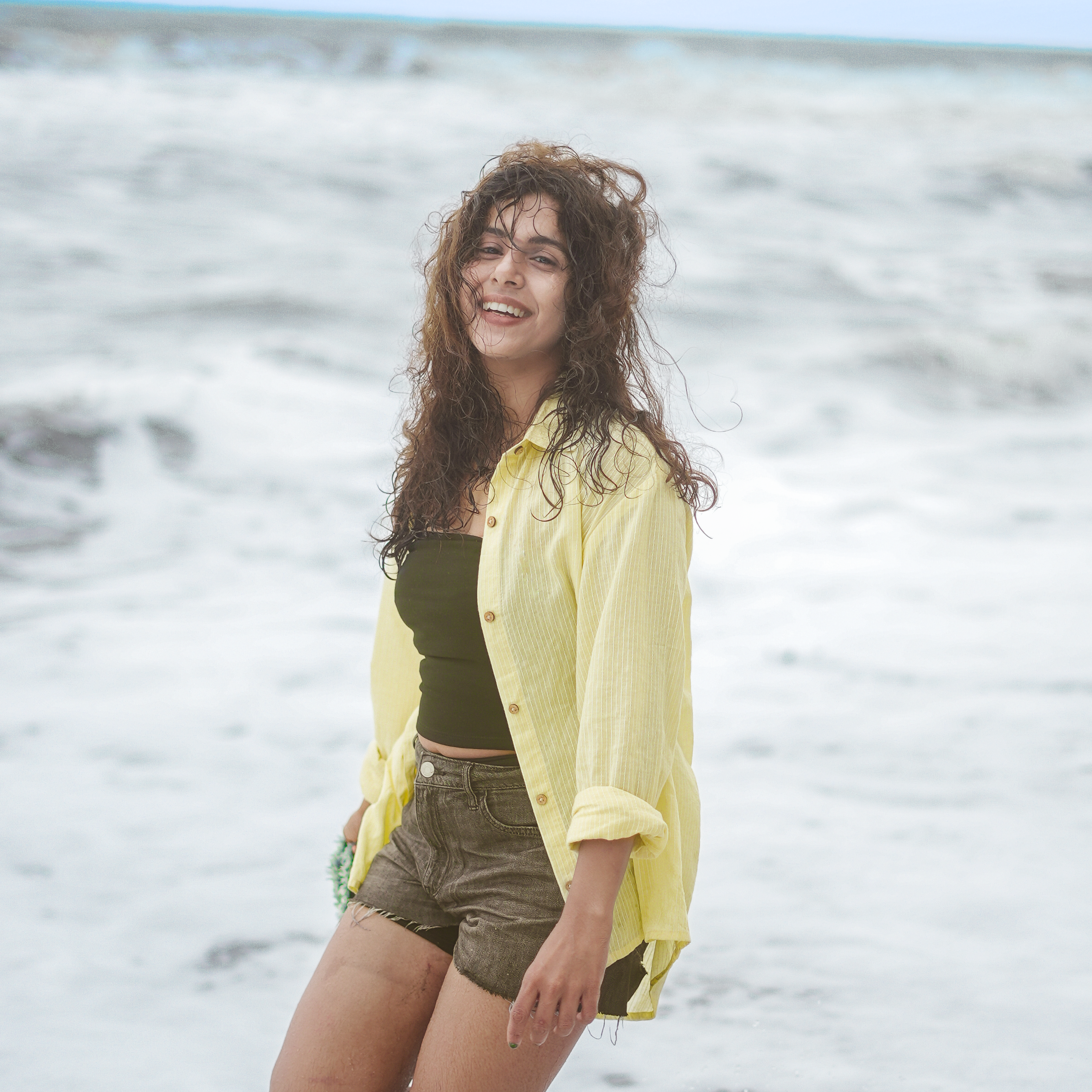 A woman standing in shallow ocean water at the beach, wearing an open light yellow button-up shirt over a dark green top and olive-brown shorts.