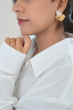 A close-up lifestyle shot of a model wearing a gold and white textured flower-shaped earring, paired with a white ribbed collared shirt and gold rings.