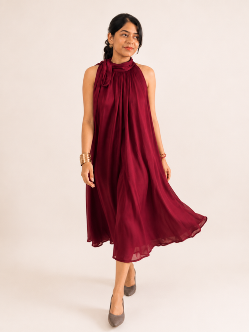A woman modeling a sleeveless, high-neck, flowy midi dress in deep burgundy, standing against a plain light beige studio background.